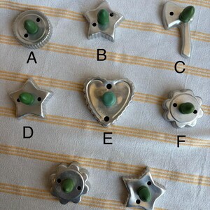 May include: A selection of eight metal cookie cutters in diverse shapes, such as stars, a heart, a flower, and a circular design. Each cutter features a green handle. The cutters are displayed on a striped surface.