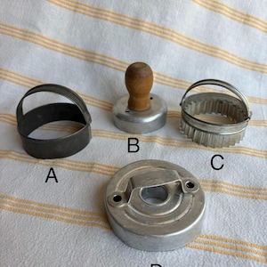 May include: Four vintage metal cookie cutters. The cutters include a round cutter with a handle, a fluted cutter with a handle, a round cutter with a wooden handle, and a round cutter with a handle.