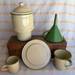 May include: A collection of vintage enamelware kitchen items in cream and green. Includes a lidded canister, a funnel, a plate, and two mugs. The items are arranged on a wooden box and a striped fabric background.