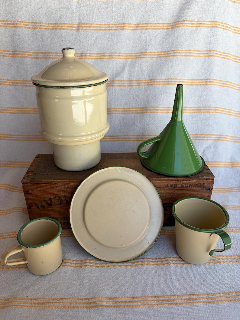 May include: A collection of vintage enamelware kitchen items in cream and green. Includes a lidded canister, a funnel, a plate, and two mugs. The items are arranged on a wooden box and a striped fabric background.