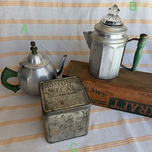 May include: An assortment of vintage tea items. Two aluminum teapots with green handles and a metal tea tin with the text "Lipton's Tea". The tin reads "The Most Delicious The World Produces". The items are on a striped fabric.