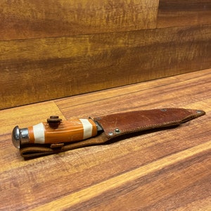 May include: A brown leather sheath with a white and orange striped handle knife. The knife has a silver metal tip and a small metal button on the handle.