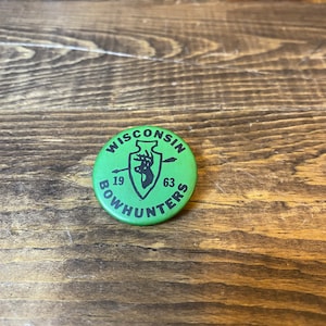 May include: Green button with the text "WISCONSIN BOWHUNTERS" in black lettering around a black silhouette of a deer with an arrow through it. The numbers "19" and "63" are on either side of the deer.