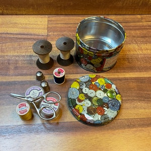 May include: A vintage sewing kit with a tin container featuring a colorful button design. The kit includes spools of thread, thimbles, and a pair of small scissors.