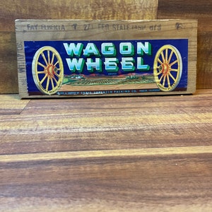 May include: A wooden crate with a blue and white label that says "Wagon Wheel" in red letters. The label also features a red, white, and blue American flag and two wagon wheels. The text on the label reads "California Quality Fruits" and "Packed & Shipped by Chris Sorensen Packing Co. Parlier, California."