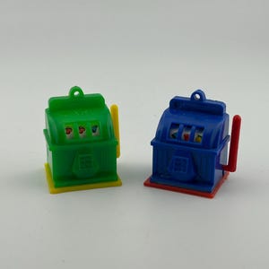 May include: Two miniature slot machine toys. One is green with a yellow handle and base, the other is blue with a red handle and base. Each has a small loop at the top.