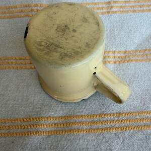 May include: A vintage, cream-colored enamelware mug with a handle. The mug shows signs of wear and tear, with dark spots and discoloration on the bottom and sides. The mug is set against a striped, off-white and yellow fabric background.
