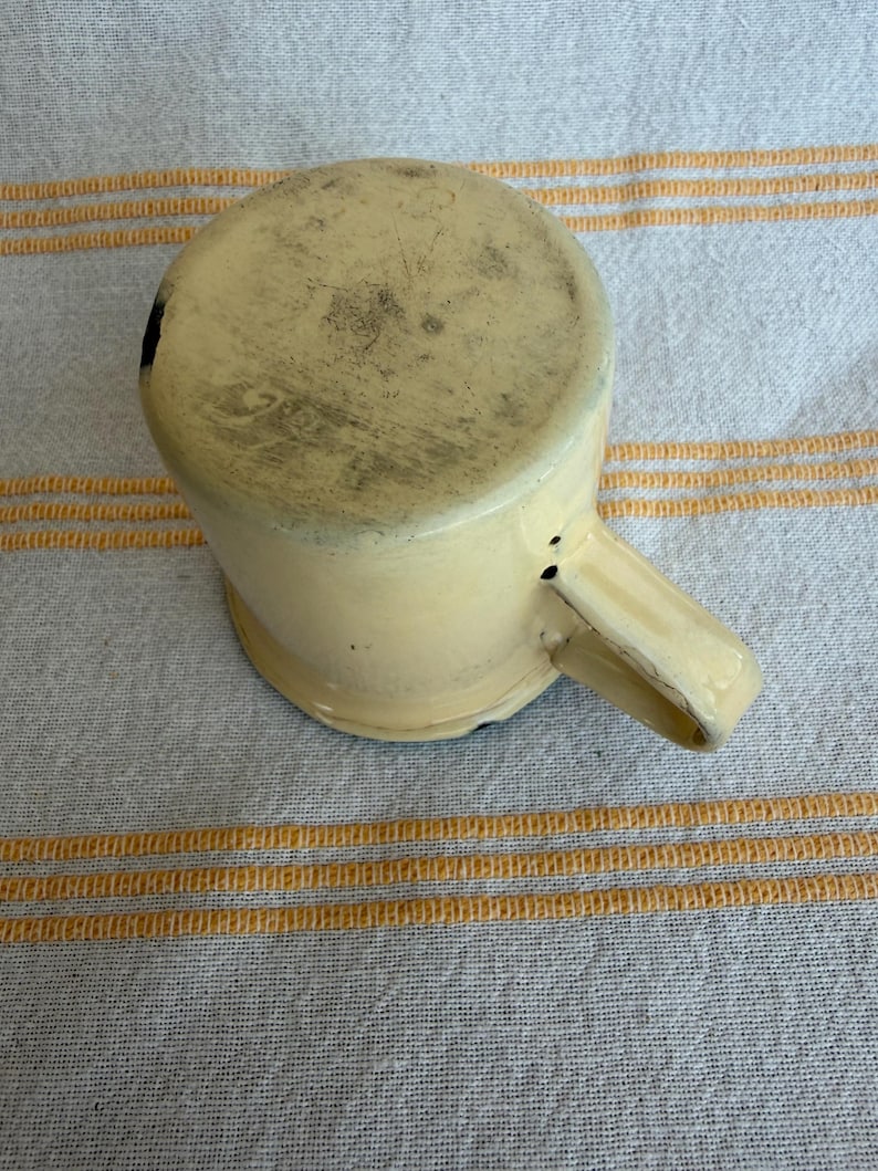 May include: A vintage, cream-colored enamelware mug with a handle. The mug shows signs of wear and tear, with dark spots and discoloration on the bottom and sides. The mug is set against a striped, off-white and yellow fabric background.