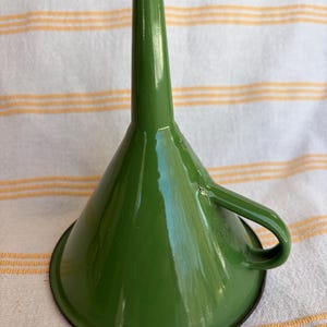 May include: A green metal funnel with a handle. The funnel has a wide base that tapers to a narrow spout. The handle is a loop shape. The funnel is painted a glossy green color with a black rim.