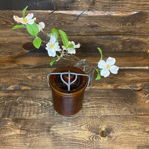 May include: A brown ceramic jar with a metal lid and handle. The jar is sitting on a wooden surface with a sprig of white flowers in a glass vase.
