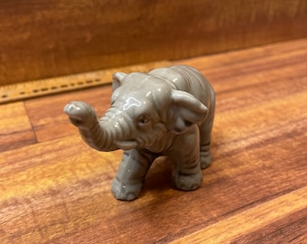 Può includere: Una piccola statuetta di elefante in ceramica grigia con la proboscide alzata. L'elefante è in piedi su una superficie di legno.