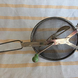 May include: An antique metal food mill with a mesh strainer and a green ceramic handle. The mill has a long handle and two curved handles on the side. The metal is tarnished, giving it a vintage look. The food mill is designed for straining and pureeing food.