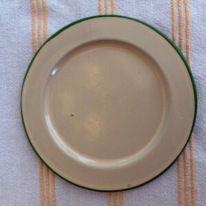 May include: A beige enamel plate with a green rim. The plate is centered on a white and yellow striped cloth. The plate is round and has a slightly raised edge.