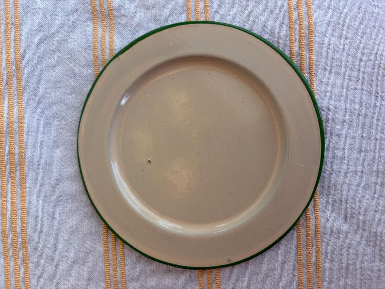 May include: A beige enamel plate with a green rim. The plate is centered on a white and yellow striped cloth. The plate is round and has a slightly raised edge.