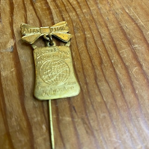 Puede incluir: Pin de color dorado con un lazo que dice "Atlas Flour" y una bolsa que dice "B. Stern & Sons Superior Wheat Flour Fancy Patent Milwaukee".