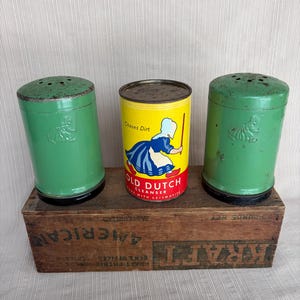 May include: An antique collection featuring two green metal salt shakers and a yellow and red Old Dutch Cleanser can. The items are displayed on a wooden Kraft box with printed text. The salt shakers have a decorative embossed design.
