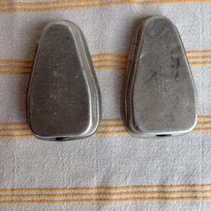 May include: Two vintage, silver-toned metal shoe stretchers, each with a rounded top and a narrow, pointed toe shape. The stretchers are slightly tarnished, showing signs of age. The background is a white cloth with yellow stripes.