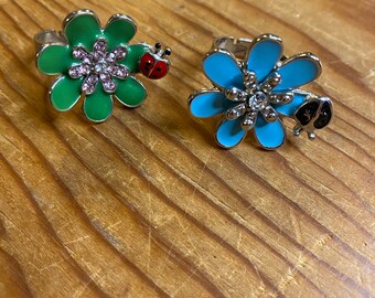 May include: Two silver rings with flower designs. One ring has a green flower with pink rhinestones and a red ladybug. The other ring has a blue flower with clear rhinestones and a black ladybug.