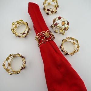 May include: A red napkin is decorated with gold-coloured napkin rings. The rings have a decorative design with red and amber-coloured gemstones. The napkin rings are arranged around the napkin on a white surface.
