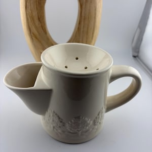 Op de afbeelding: Een crèmekleurige scheerkom met een handvat en een zijkom. De kom heeft een decoratief reliëfpatroon en een geperforeerde bovenkant. Op de achtergrond staat een houten sculptuur met een ovale uitsparing.