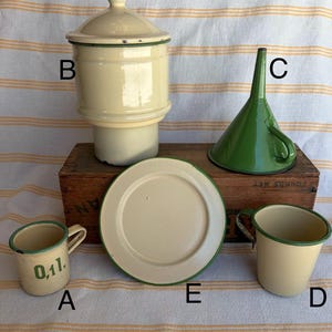 May include: A collection of vintage enamelware kitchen items. Includes a cream-colored canister with a lid, a green funnel, a small mug with "0,1l" text, a plate, and a small cream-colored cup, all with green trim. The items are arranged on a wooden box.