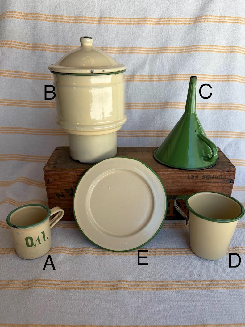 May include: A collection of vintage enamelware kitchen items. Includes a cream-colored canister with a lid, a green funnel, a small mug with "0,1l" text, a plate, and a small cream-colored cup, all with green trim. The items are arranged on a wooden box.