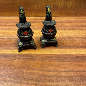Puede incluir: Dos saleros y pimenteros de hierro fundido pintados de negro con forma de estufas de leña con corazones rojos y detalles florales.
