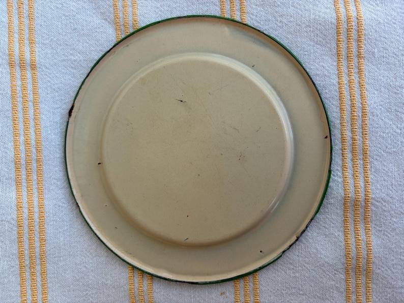 May include: A vintage, off-white enamel plate with a green rim. The plate has a slightly raised center and shows signs of wear, adding to its antique charm. The plate is set against a striped background.