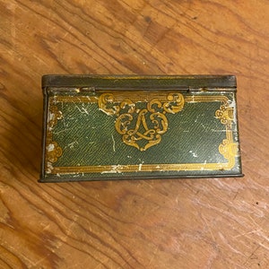 Puede incluir: Una caja de metal vintage de color verde con ribetes dorados y un diseño decorativo. La caja tiene una tapa con bisagras y está decorada con un patrón floral dorado y una letra "L" en el centro.