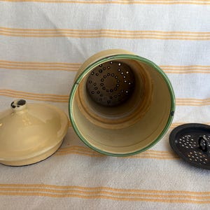 May include: A vintage, cream-colored ceramic coffee pot with a matching lid and a separate, perforated insert. The rim of the pot is accented with a green stripe. The pot is open, revealing the interior and the insert.