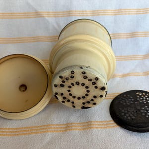 May include: An antique ivory-colored spice grinder with a matching lid and a separate dark brown perforated lid. The grinder has a cylindrical body with a textured base and a circular top. The lid is round and flat.