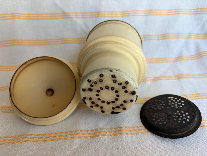 May include: An antique ivory-colored spice grinder with a matching lid and a separate dark brown perforated lid. The grinder has a cylindrical body with a textured base and a circular top. The lid is round and flat.