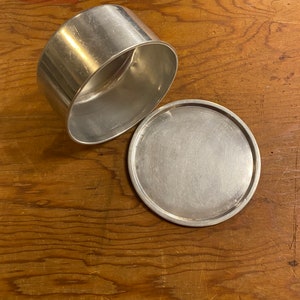 May include: A silver metal container with a lid. The container is round and has a smooth surface. The lid is also round and has a smooth surface.