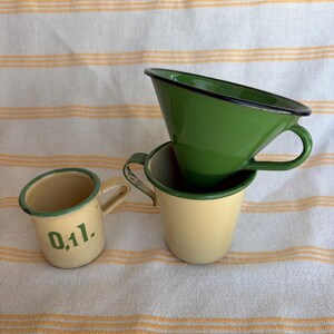 May include: Three vintage enamelware cups in shades of green and cream. One cup has the text "0,1l." in green. The cups have black rims and small handles. The cups are stacked on a white and yellow striped surface.