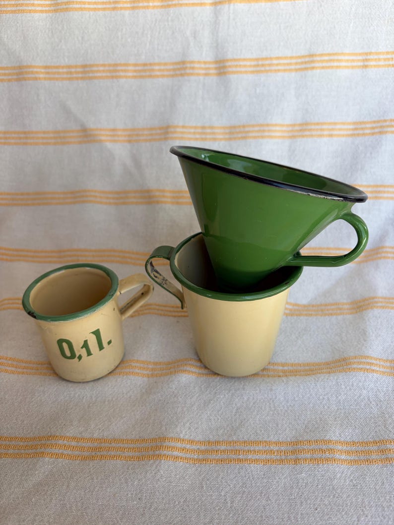 May include: Three vintage enamelware cups in shades of green and cream. One cup has the text "0,1l." in green. The cups have black rims and small handles. The cups are stacked on a white and yellow striped surface.