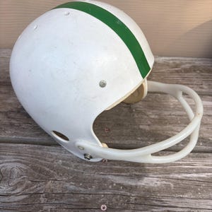 Puede incluir: Un casco de fútbol americano blanco con una franja verde central que recorre desde la parte delantera hasta la trasera. El casco incluye una máscara facial de plástico blanco sujeta con remaches metálicos y orificios de ventilación.