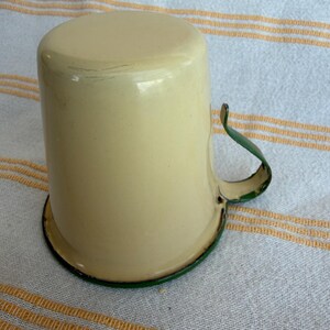 May include: Cream-colored enamel mug with a green handle and rim. The mug has a cylindrical shape and a slightly rounded top. The handle is curved and attached to the side of the mug. The mug sits on a white and yellow striped surface.