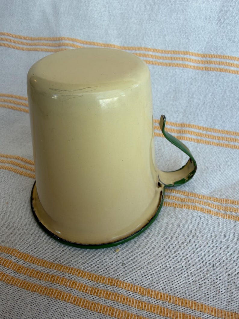 May include: Cream-colored enamel mug with a green handle and rim. The mug has a cylindrical shape and a slightly rounded top. The handle is curved and attached to the side of the mug. The mug sits on a white and yellow striped surface.