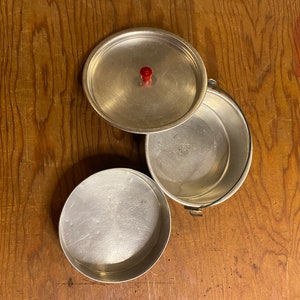 May include: A set of three silver aluminum camping cookware. The set includes a pot with a lid and a smaller bowl. The lid has a red knob.