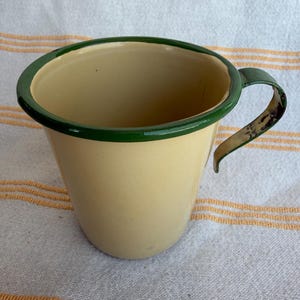 May include: A vintage enamelware mug with a cream-colored body and a dark green rim and handle. The mug has a classic, rounded shape and is likely used for drinking beverages. The mug is sitting on a striped fabric.