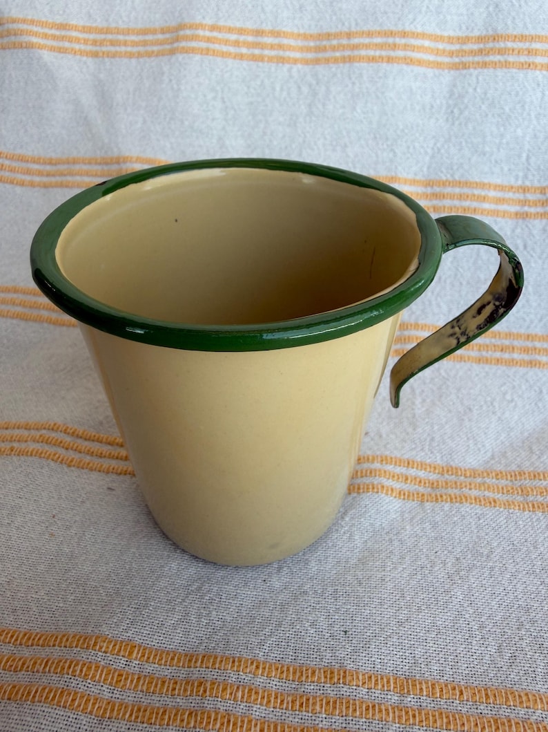 May include: A vintage enamelware mug with a cream-colored body and a dark green rim and handle. The mug has a classic, rounded shape and is likely used for drinking beverages. The mug is sitting on a striped fabric.