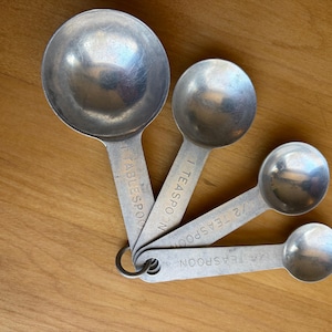 May include: A set of four stainless steel measuring spoons, including 1 tablespoon, 1 teaspoon, 1/2 teaspoon, and 1/4 teaspoon. The spoons are attached to a metal ring and are arranged on a wooden surface.