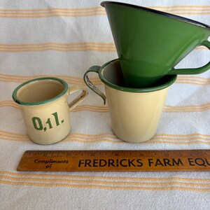 May include: Three vintage enamelware cups in cream and green, with black trim. One cup has the number "0,1l." printed on it. A wooden ruler with the text "FREDRICKS FARM EQU" is also present.