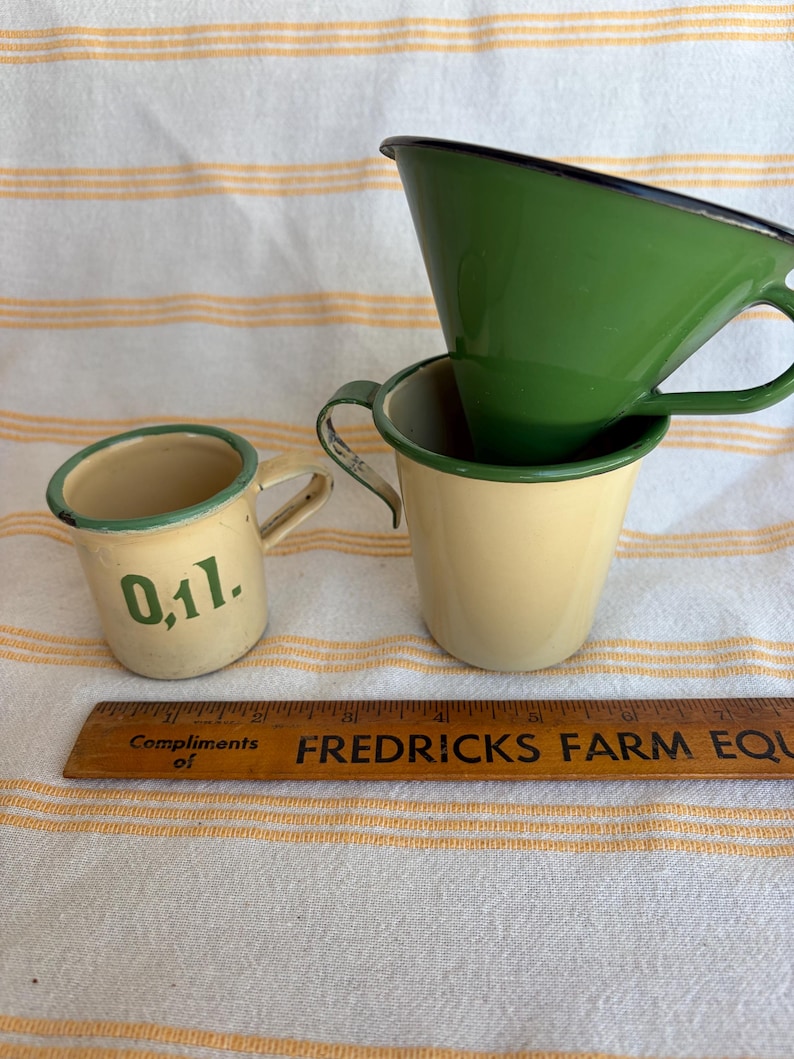 May include: Three vintage enamelware cups in cream and green, with black trim. One cup has the number "0,1l." printed on it. A wooden ruler with the text "FREDRICKS FARM EQU" is also present.