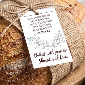Könnte beinhalten: Ein frisch gebackenes Brot, in Jute gewickelt und mit Bindfaden gebunden, liegt auf einem Holzschneidebrett. Ein Anhänger liest: "Sie brachen Brot...Apostelgeschichte 2:46" und "Gebacken mit Absicht, Geteilt mit Liebe."
