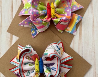 May include: Two colorful hair bows with crayon embellishments. One bow features a vibrant mix of patterns and colors, including polka dots and school-themed designs. The other bow has a white background with school supplies and red trim.