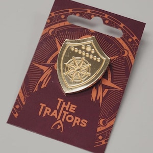 May include: A gold-colored shield-shaped pin featuring a compass rose design and small circles. The pin is on a maroon card with the text "The Traitors" and a celestial design.