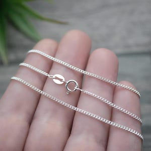 May include: A thin, silver-coloured curb chain necklace featuring small, interlocking oval links. The chain includes a round spring ring clasp and a small, flat, oval-shaped metal tag attached near the closure mechanism for secure fastening.