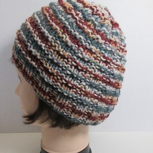 May include: A hand-knitted beanie hat in a mix of colors including teal, burgundy, cream, and tan. The hat is made with a textured stitch pattern and is displayed on a mannequin head. The hat is a winter accessory.