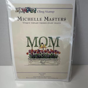 May include: A cling stamp package featuring a floral design with the word "MOM" in green, leafy letters. Below the letters is a planter box filled with red and purple flowers. The stamp is from Michelle Masters.
